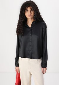 Chemise noire en satin à manches longues avec un col à boutons et des boutons sur le devant, associée à un pantalon de couleur claire et un sac à main rouge.