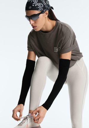 Oseba s sončnimi očali, črno rutko, sivo srajco z zavihanimi rokavi, črnimi rokavi za roke in svetlimi legicami, ki veže vezalke na belih supergah.