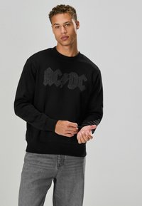 Fekete pulóver, melyen egy texturált AC/DC logó látható ezüst színben, kerek nyakkivágással, bordázott mandzsettákkal és aljjal, szürke farmernadrággal párosítva.