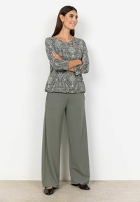 Haut à manches longues en tissu gris et vert à motifs, associé à un pantalon olive à taille haute et jambes larges, avec des chaussures noires.