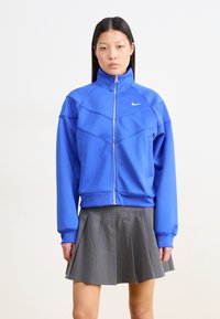Blå zip-up jakke med høy krage, som har sporty sømmer og en liten hvit logo. Kombinert med et grått plissékjørt.