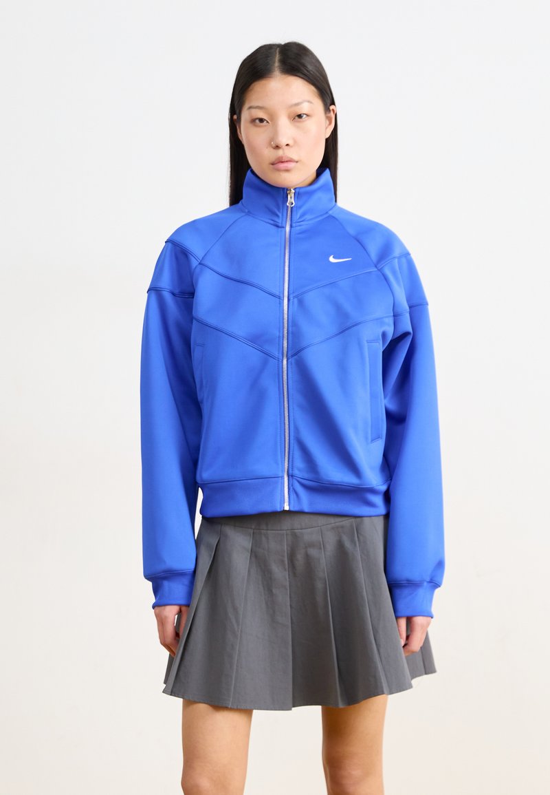 Blå zip-up jakke med høy krage, som har sporty sømmer og en liten hvit logo. Kombinert med et grått plissékjørt.