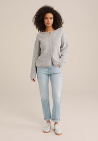 Grå stickad cardigan med rund halsringning och framknappar, i kombination med ljusblå jeans och sneakers. Har långa ärmar och en avslappnad passform.