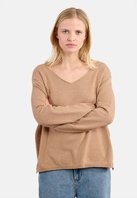 Donna bionda con espressione neutra, braccia incrociate, che indossa un maglione beige largo e jeans blu, in piedi di fronte a uno sfondo chiaro e uniforme.