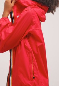 Czerwona, lekka kurtka przeciwdeszczowa z kapturem, z zamkiem z przodu, bocznymi kieszeniami i kontrastowymi paskami wzdłuż krawędzi zamka.