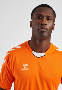 Orange sportsjersey med V-hals, hvide accenter på skuldrene, og et logo på brystet. Lavet af letvægts, åndbart materiale.