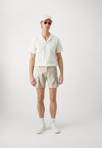 Camisa de botones de manga corta en un color beige claro con rayas texturizadas, combinada con pantalones cortos de cargo color caqui claro y zapatillas blancas. Incluye una gorra blanca y gafas de sol.