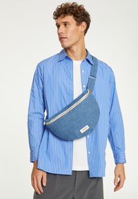 Sac en denim en forme arrondie, avec une fermeture éclair dorée et une petite étiquette. Porté en bandoulière sur une chemise à rayures bleues et un pantalon gris.