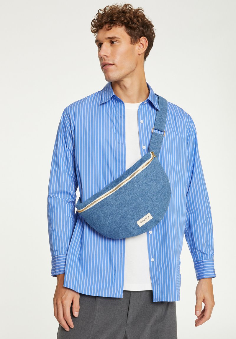 Sac en denim en forme arrondie, avec une fermeture éclair dorée et une petite étiquette. Porté en bandoulière sur une chemise à rayures bleues et un pantalon gris.