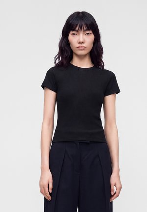 Jeune femme aux cheveux noirs ondulés porte un haut court noir ajusté et un pantalon large foncé, debout devant un fond clair uni.