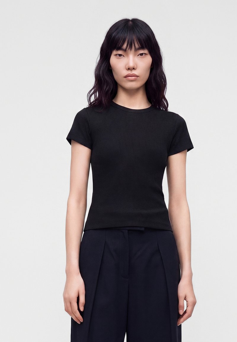 Jeune femme aux cheveux noirs ondulés porte un haut court noir ajusté et un pantalon large foncé, debout devant un fond clair uni.