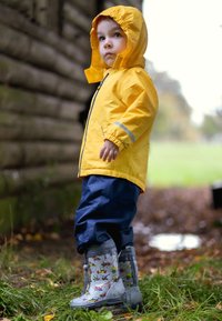 Geel waterdicht jack met een capuchon, blauwe broek en grijze regenlaarzen met patronen van bouwvoertuigen, staande op gras.