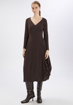 Abito a maniche lunghe con scollatura a V profonda in tessuto marrone scuro, caratterizzato da una silhouette fluida con un dettaglio drappeggiato laterale. Abbinato a stivali neri al ginocchio.