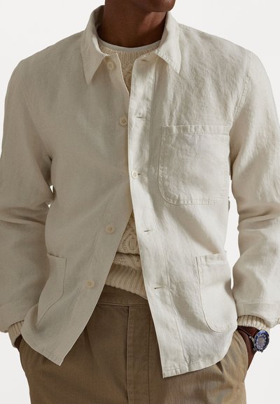 Light beige linen shirt with a button-up front, long sleeves, and a chest pocket, paired with a textured sweater underneath.
