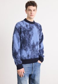 Kék és tengerészkék pulóver textured mintával, kerek nyakkivágással és bordás mandzsettával, világoskék farmerral párosítva.