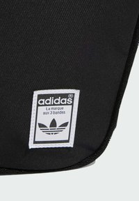 Čierna látková taška s bielou nášivkou loga Adidas a trojpruhovým dizajnom, proti textúrovanému pozadiu so šikmým pruhovaním.