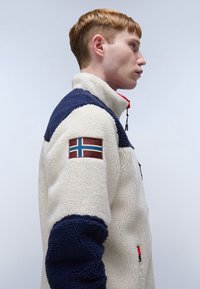 Fleece jacket in cream and navy, featuring a high collar, zipper, and a patch with a flag. Textured fabric with contrasting sleeves.