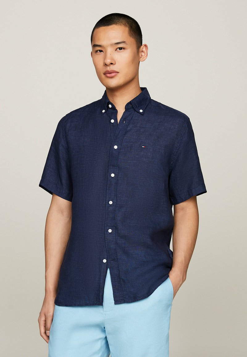 Camicia blu navy a maniche corte con bottoni, realizzata in un tessuto texturizzato, dotata di colletto e piccolo logo sul petto. Abbinata a pantaloni azzurri chiari.