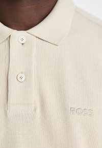 Camisa polo beige com riscas, com uma pala de dois botões, apresentando "BOSS" bordado na mesma cor no lado esquerdo. Material em mistura de algodão.