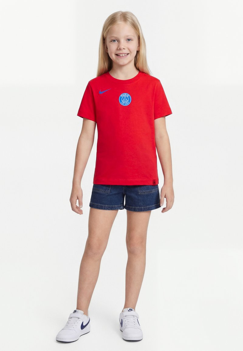 Junges Mädchen mit langen blonden Haaren, das ein rotes Paris Saint-Germain T-Shirt, blaue Jeans-Shorts und weiße Nike-Sneaker trägt und lächelt.