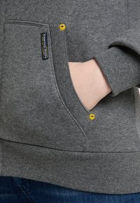 Sudadera gris con puños y dobladillo acanalados. Presenta un bolsillo frontal con botones de acento amarillos y una etiqueta en el lateral. Textura suave.