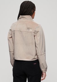 Light beige cropped jacket with a high collar, made of soft material, featuring an embossed logo on the upper back, and elastic cuffs.