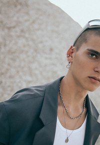 Grå blazer, vit tanktop, lager av silverkedjor, silverörhänge med fredssymbol och solglasögon. Släta texturer med subtila detaljer.
