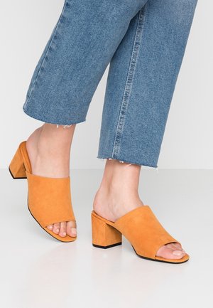 Persoon draagt blauwe cropped jeans en oranje suède open-teen blokhakken, staand op een wit oppervlak.