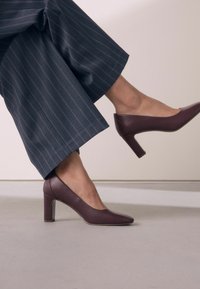Burgundy nahka korkokengät, joissa on terävä kärki ja laatikkokorko, yhdistettynä tummansinisiin pinstripe-laaraluisiin housuihin. Silkkinen pinta ja tyylikäs muotoilu.