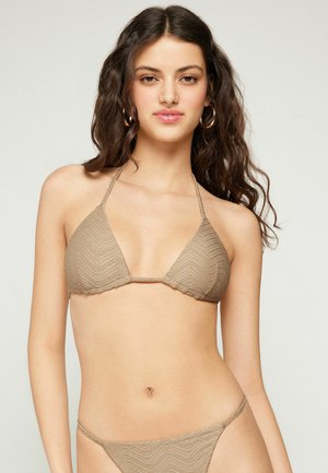 Mujer con pelo largo y oscuro, vestida con parte superior e inferior de bikini beige texturizado, de pie frente a un fondo claro y liso.