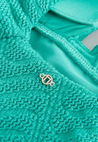 Tissu turquoise avec motif tricot ondulé texturé, sections en mesh lisses et petit logo métallique rond doré fixé près de la couture.
