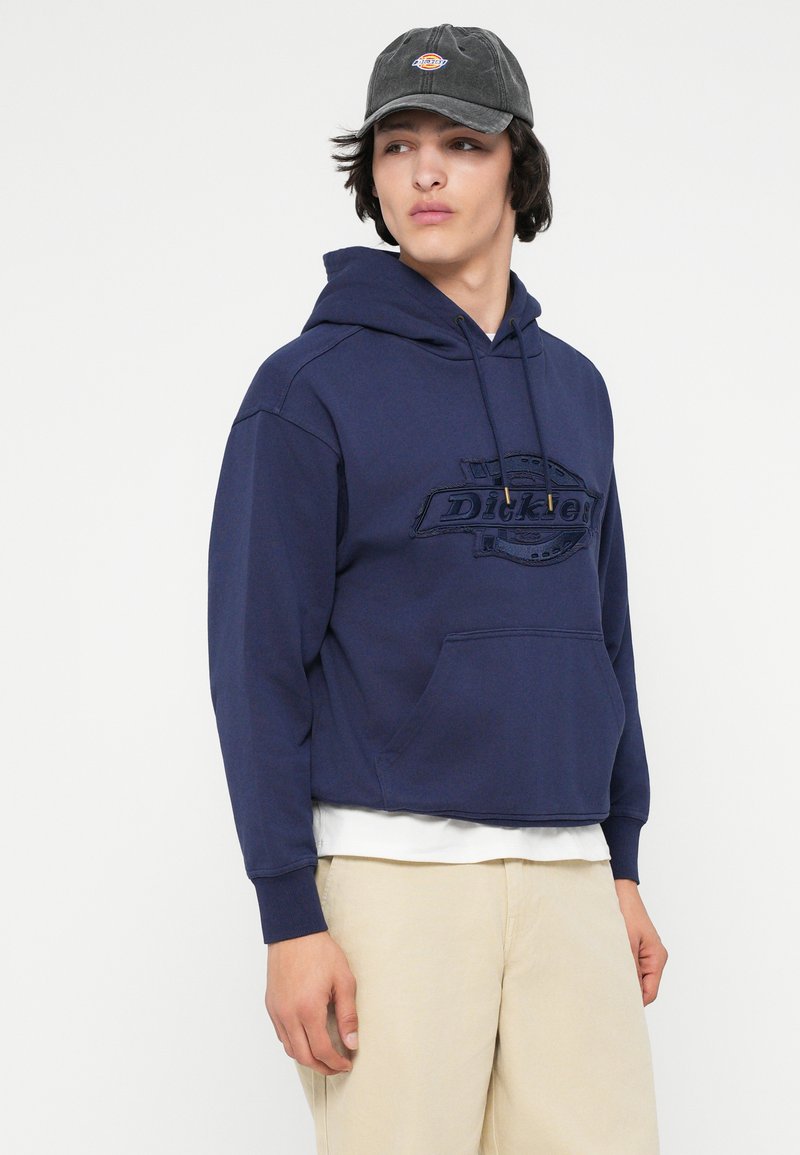 Navyblauwe hoodie met een logo op de voorkant, voorzien van een capuchon met trekkoord en een voorkantvak. Draag ik over een wit shirt en gecombineerd met beige broek.