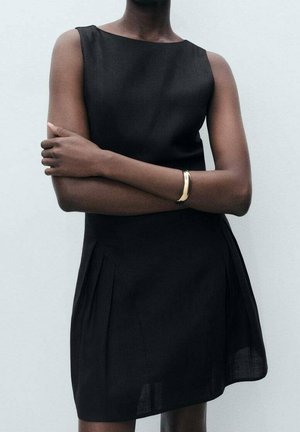 Persona con vestido negro sin mangas y los brazos cruzados, de pie contra un fondo claro y sencillo, con una pulsera dorada en la muñeca izquierda.