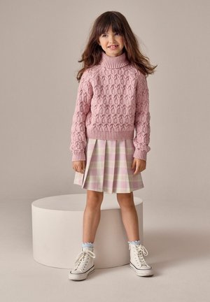 Pige med langt brunt hår iført en lyserød strikket sweater, lyserødt ternet plisseret nederdel, hvide høje sneakers, stående i et neutralt studie miljø.