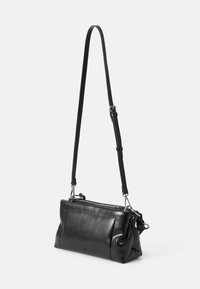 Sac à bandoulière en cuir noir avec bandoulière réglable, ferrures en argent et fermeture éclair, présenté suspendu sur un fond blanc.
