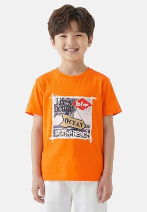 Garçon souriant portant un t-shirt orange avec le texte « Life's better OCEAN at the beach » et un short blanc, devant un fond uni.