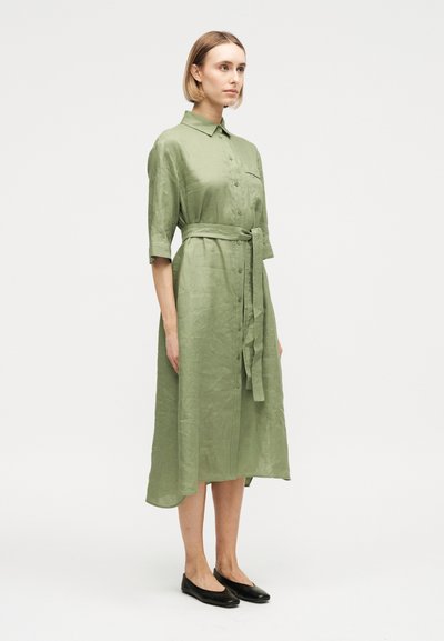 Γυναίκα με ανοιχτό πράσινο, κουμπωτό, midi φόρεμα με ζώνη και μανίκια μέχρι τον αγκώνα, φορόντας μαύρα παπούτσια χωρίς τακούνι, στέκεται μπροστά σε απλό φόντο.