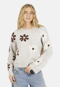 Pull en tricot beige clair avec des motifs floraux marron et blanc, manches côtelées et col rond, porté sur un jean bleu clair.
