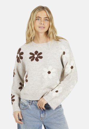 Lichtbeige gebreide trui met bruine en witte bloemenpatronen, geribbelde manchetten en een ronde halslijn, gedragen over lichtblauwe jeans.