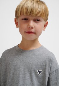 Grå bomulds t-shirt med rund hals og korte ærmer, med et blåt og hvidt logo-lap på venstre bryst. Blød tekstur.