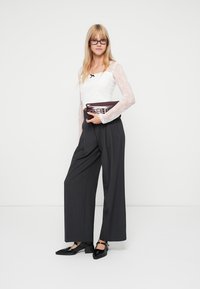 Bela čipkasta bluza z dolgimi rokavi, črne hlače širokega kroja z visokim pasom in elegantni črni čevlji. Drži bordo clutch z dekorativnimi poudarki.