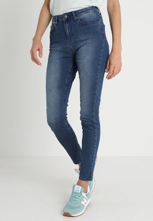 Niebieskie dżinsy typu slim-fit noszone z jasnoniebieskimi i białymi trampkami, pokazane od połowy tułowia do stóp na jednolitym tle.