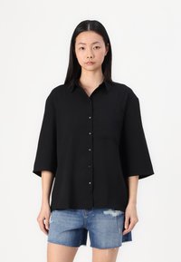 Black button-up shirt with a collar, short sleeves, and a single chest pocket. Worn over blue denim shorts, featuring a distressed hem.