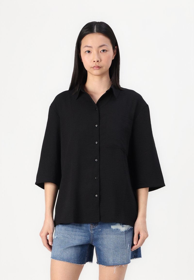 Black button-up shirt with a collar, short sleeves, and a single chest pocket. Worn over blue denim shorts, featuring a distressed hem.