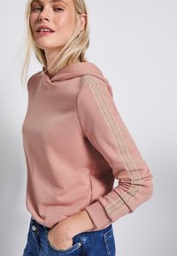 Sudadera con capucha rosa de textura suave, con rayas deportivas beige en las mangas. La lleva puesta con vaqueros azules, con las manos en las caderas.