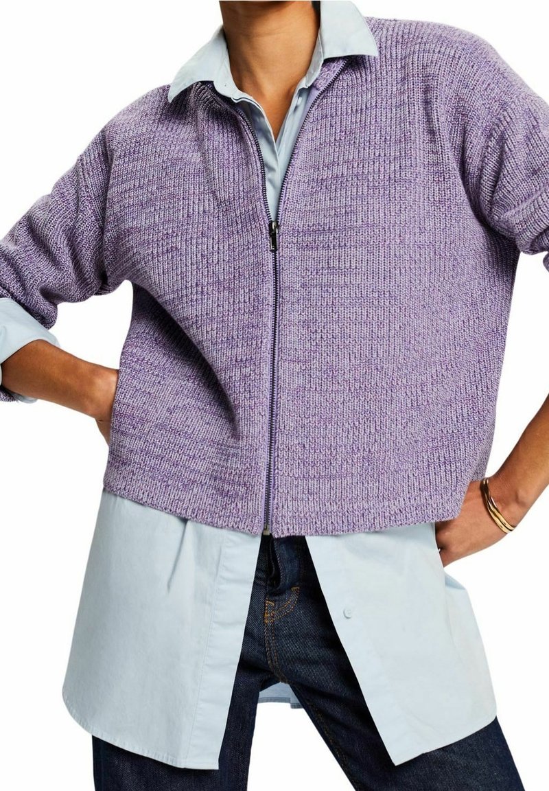 Personne portant une chemise longue bleu clair sous un pull court en tricot lavande à fermeture éclair, associé à un jean bleu foncé et un bracelet en or.
