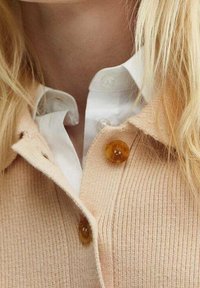 Beige ribbed knit cardigan with amber buttons, layered over a white shirt. The fabric appears soft and structured, featuring a collar detail.