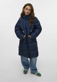 Granatowa kurtka puchowa z kapturem, z kieszeniami na zamek i pikowanym wzorem. Noszona na luźno dopasowanych jasnych dżinsach i zielonych trampkach.