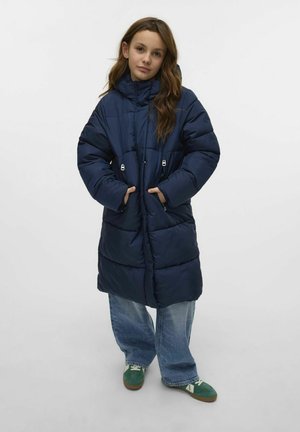 Giacca piumino blu navy con cappuccio, tasche con zip e design trapuntato. Indossata sopra jeans in denim chiaro a vestibilità ampia e sneakers verdi.