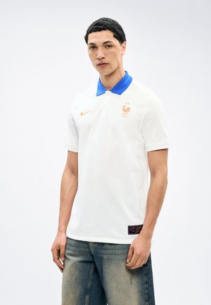 Jonge man in een wit poloshirt met blauwe kraag en goudkleurig haan-embleem en verweerde blauwe spijkerbroek, staand tegen een witte achtergrond.